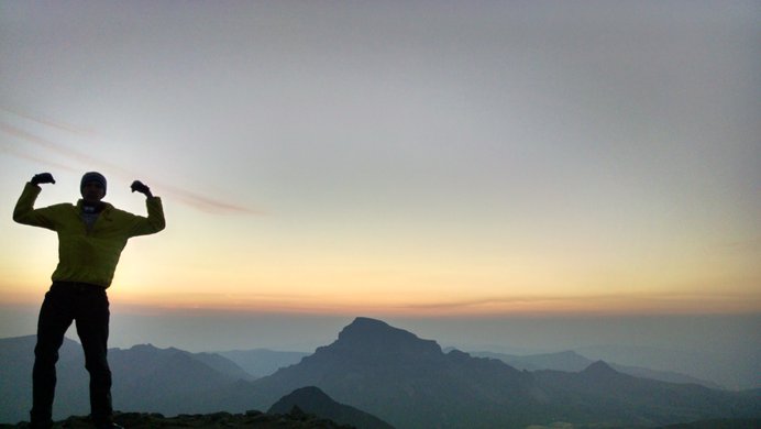 A hiker atop Wetterhorn Peak flexing his arms silhouetted against the sunrise with Uncompahgre Peak beyond